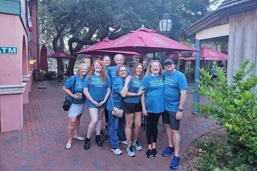 A group of people standing under a red umbrella in an outdoor setting with brick pavement and greenery. 5