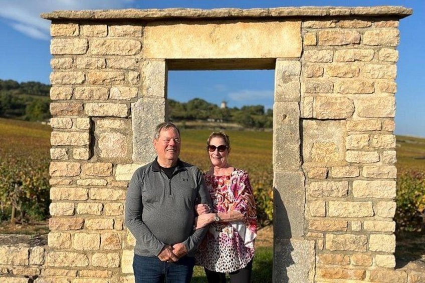 Twee personen staan voor een stenen gebouw, met een heldere lucht en velden op de achtergrond. 5