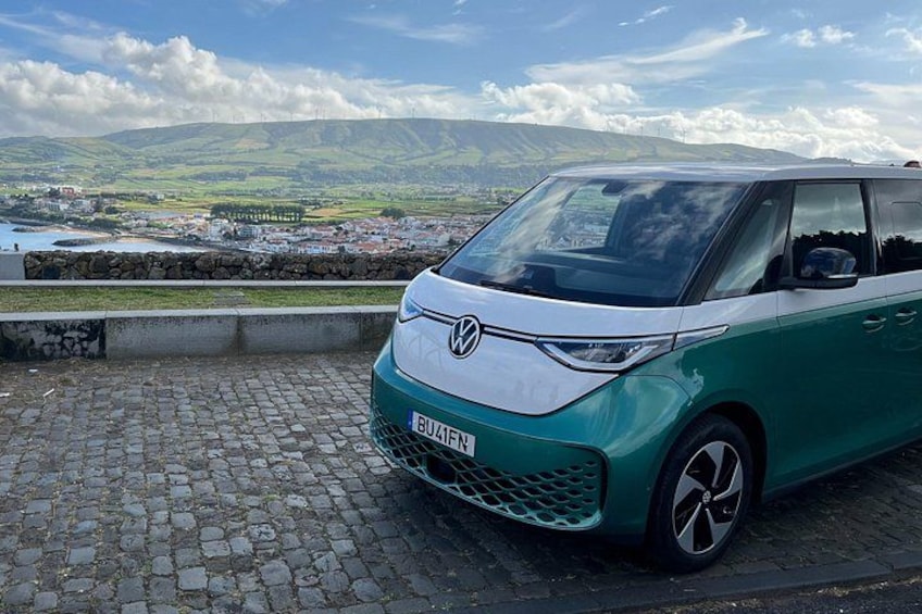 Un coche verde y blanco estacionado en una calle adoquinada, con vistas panorámicas de colinas y un cuerpo de agua al fondo. 1