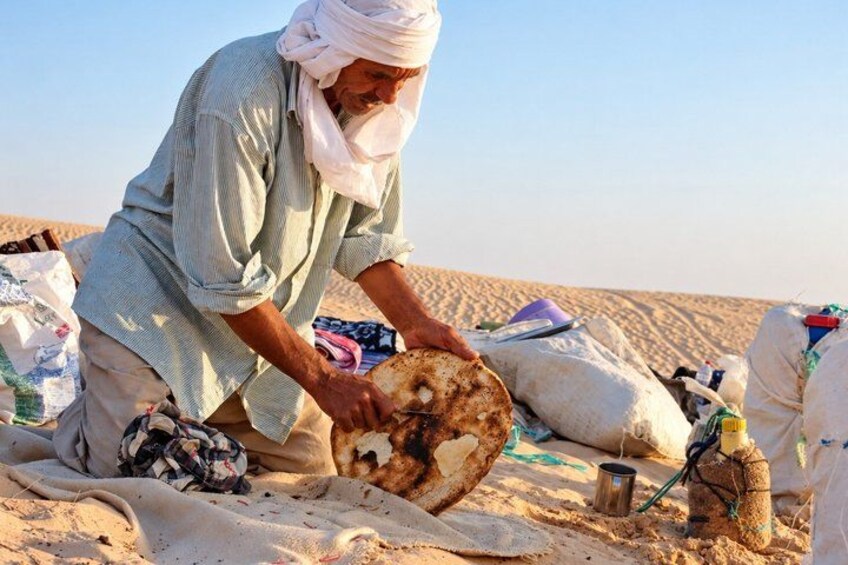 A person kneeling on sand, handling a large, round object, possibly a bread or pastry, with bags and a metal container nearby. 4