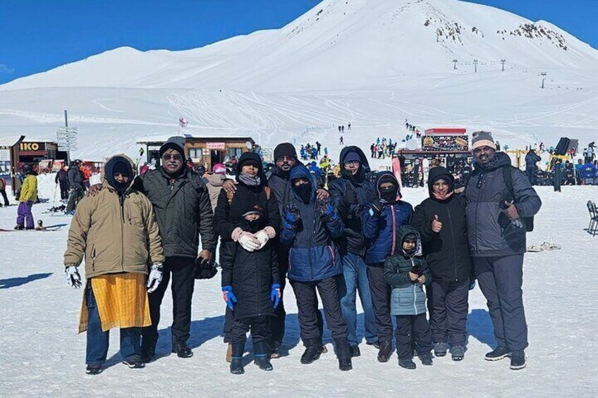 A group of people standing on a snowy mountain with ski lifts and a ski resort in the background. 3