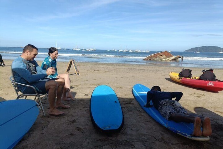 A beach with kayaks and surfboards, people sitting and relaxing, and boats in the water. 1