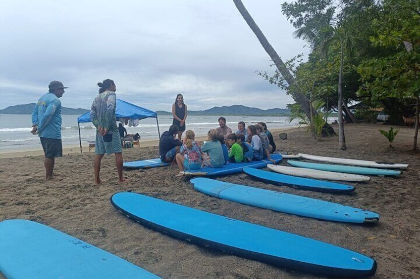 A group of people sitting on the beach under a blue tent, with surfboards lined up in the sand. 2