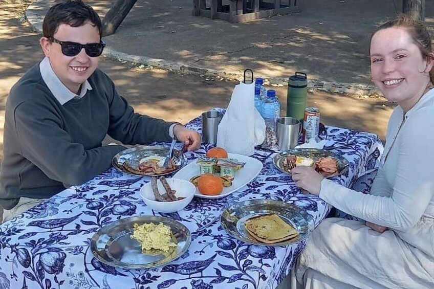 Two people dining at an outdoor table with a blue and white patterned tablecloth, plates of food, and various drink containers. 2