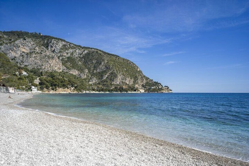 A pebble beach with clear water, a rocky hillside, and a distant building. 5