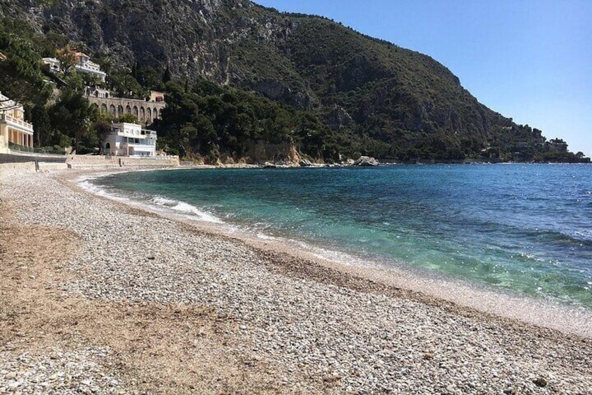 A pebble beach with clear water, a rocky hillside, and buildings on the left. 4
