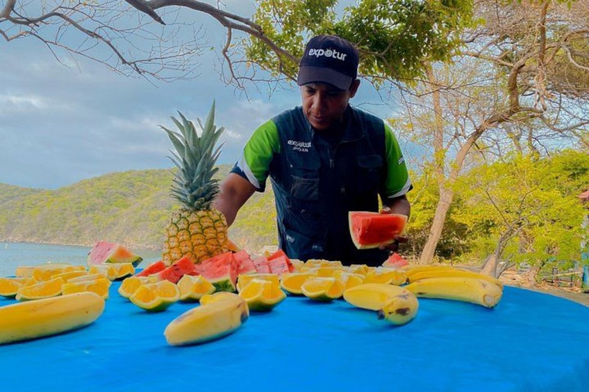 Una persona de pie junto a una mesa con diversas frutas, como plátanos, sandía y piña. 3
