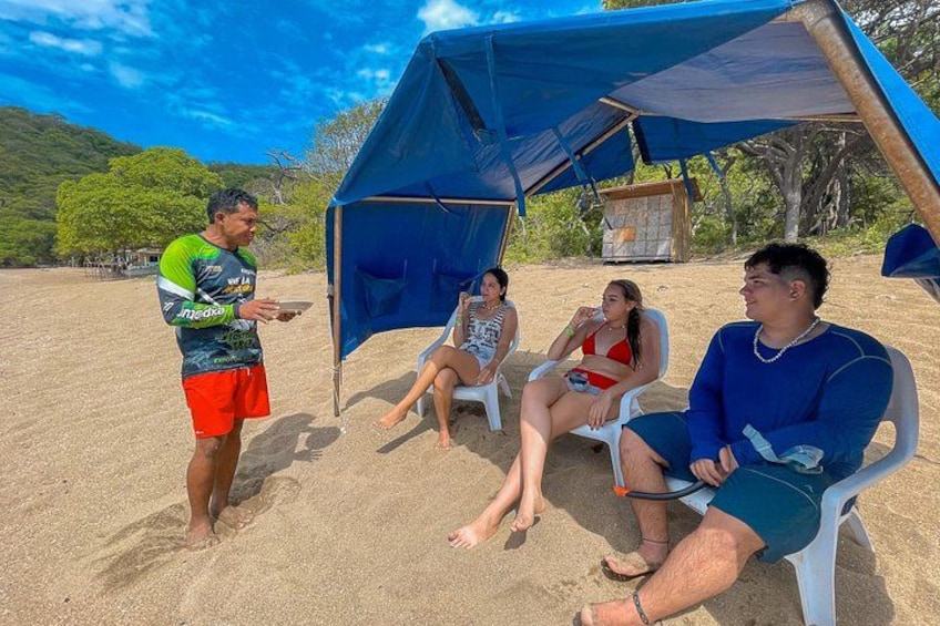 Escena de playa con gente sentada bajo una carpa azul, arena y árboles al fondo. 2