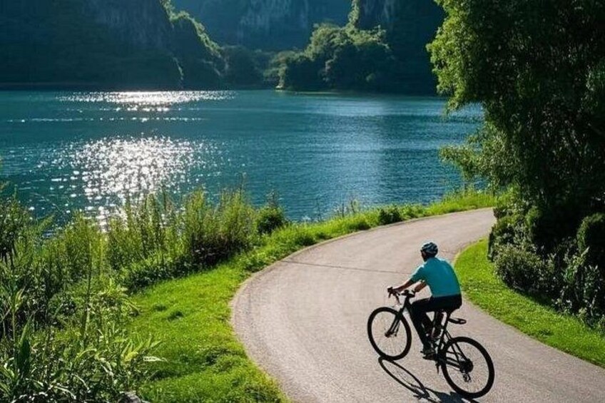 En cyklist på en asfalteret sti ved siden af en sø med grøn vegetation og bakker i baggrunden. 5