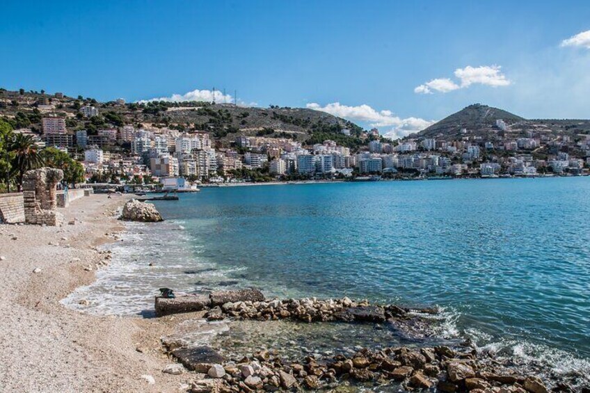 A coastal cityscape with a beach, clear blue waters, and buildings on a hillside. 1