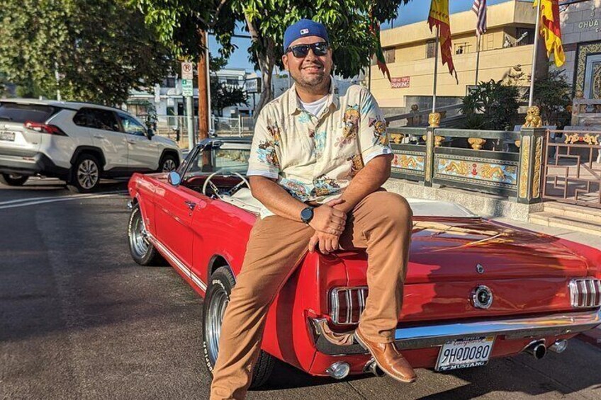 A man sitting on a red vintage car with a license plate, parked on a street with other cars and buildings in the background. 5