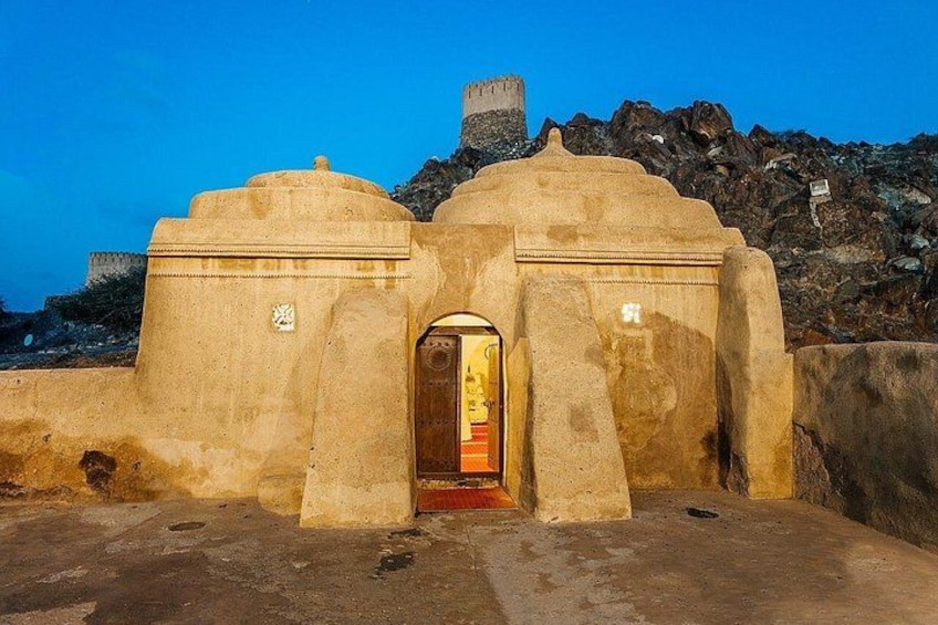 A historical building with a central archway, a stone wall, and a rocky hill in the background. 1