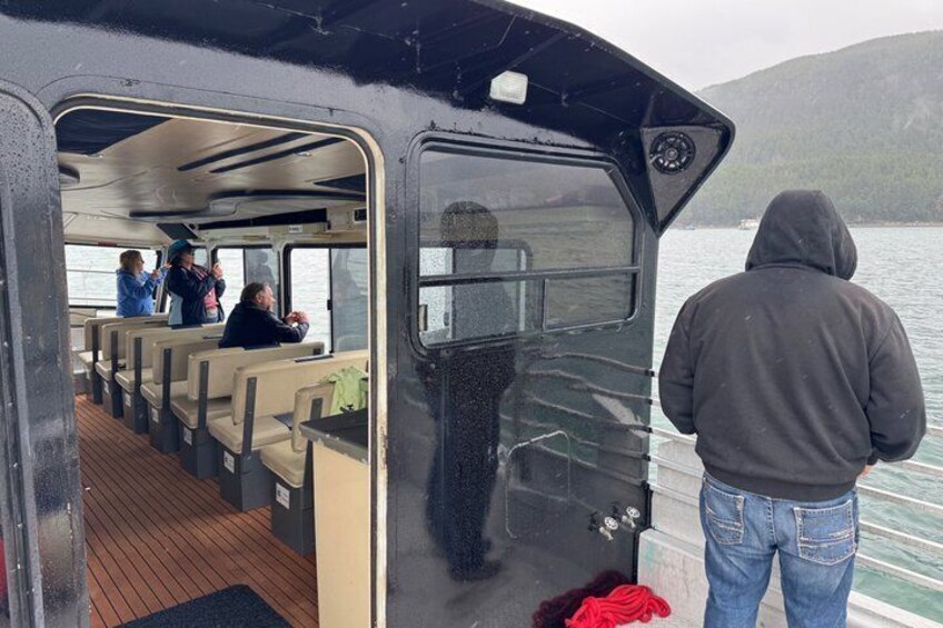 Interior de un barco con pasajeros, una persona de pie en la proa y una colina boscosa al fondo. 4