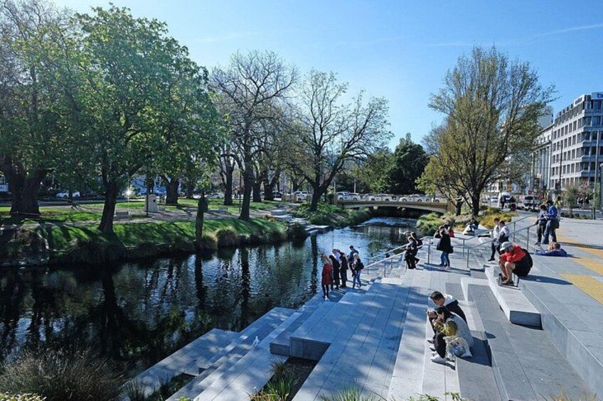 Un parque junto al río con gente caminando y sentada en bancas, árboles y un puente al fondo. 2