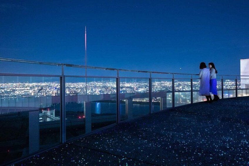 A nighttime cityscape viewed from a high vantage point with a glass railing. 5