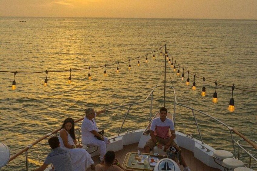 A boat with people sitting and standing, decorated with string lights, on a body of water during sunset. 2