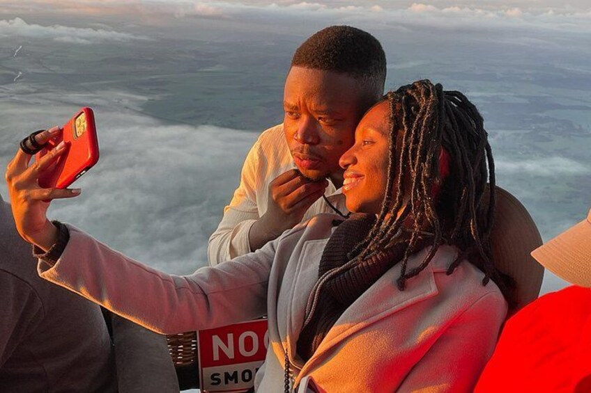 A person taking a selfie with a smartphone, another person in the background, and a sign with 'NO SMOKING' visible. 4