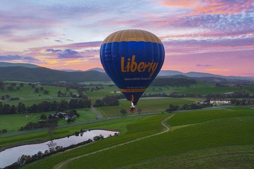 A hot air balloon with 'Liberty Balloon Flights' written on it, flying over a rural landscape with fields and a small body of water. 1