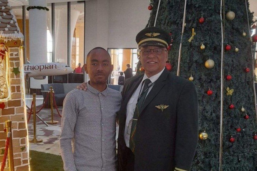 Two men in a decorated indoor setting with a Christmas tree and a model airplane. 5