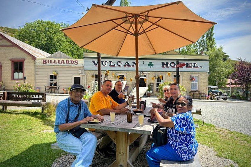 A group of people sitting at an outdoor table with a large umbrella, in front of a building labeled 'Billiards' and 'Vulcan Hotel.' 5