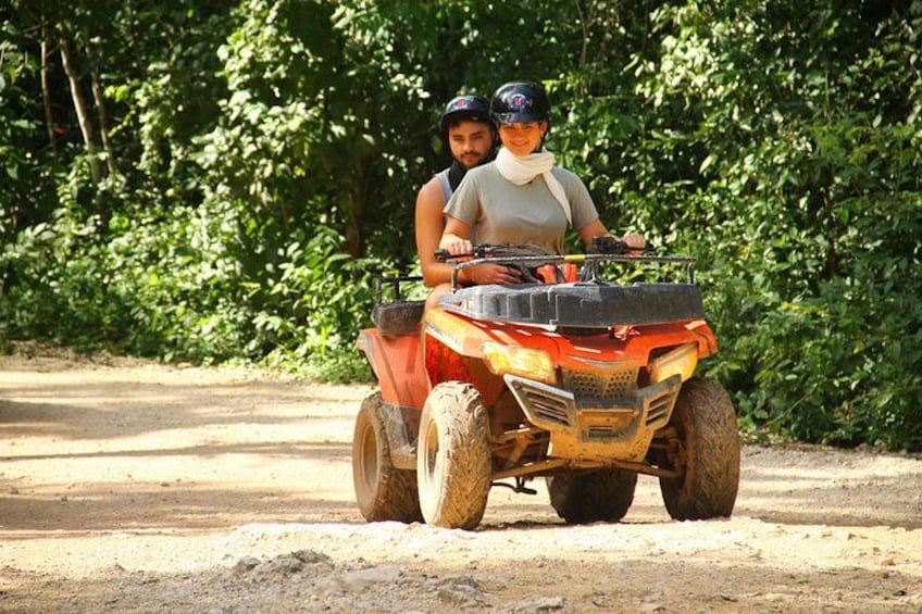 Two people on an orange ATV on a dirt road with dense green foliage in the background. 1