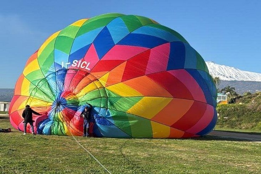 A colorful hot air balloon with the text 'K-SICL' on it, being inflated on a grassy field with a mountain in the background. 2