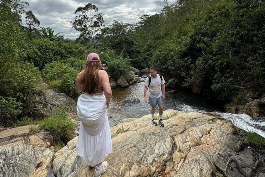 Two people standing on rocky terrain near a flowing river, surrounded by dense greenery. 1