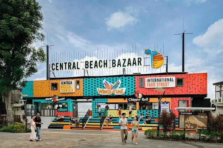 Een kleurrijk gebouw met borden voor Central Beach Bazaar, Sentosa Musical Theatre, Wings Over Time, en Chipper Roll. 4