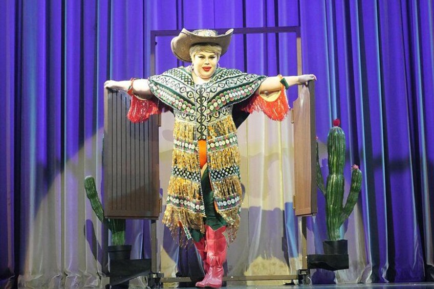 Une personne vêtue d’un costume mexicain traditionnel, coiffée d’un sombrero, se tient sur une scène décorée de cactus et surmontée d’un rideau bleu. 4