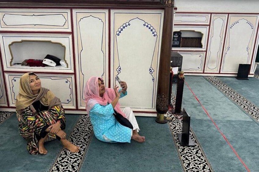 Two women sitting on patterned rugs in a room with ornate wall decorations and a wooden column. 4