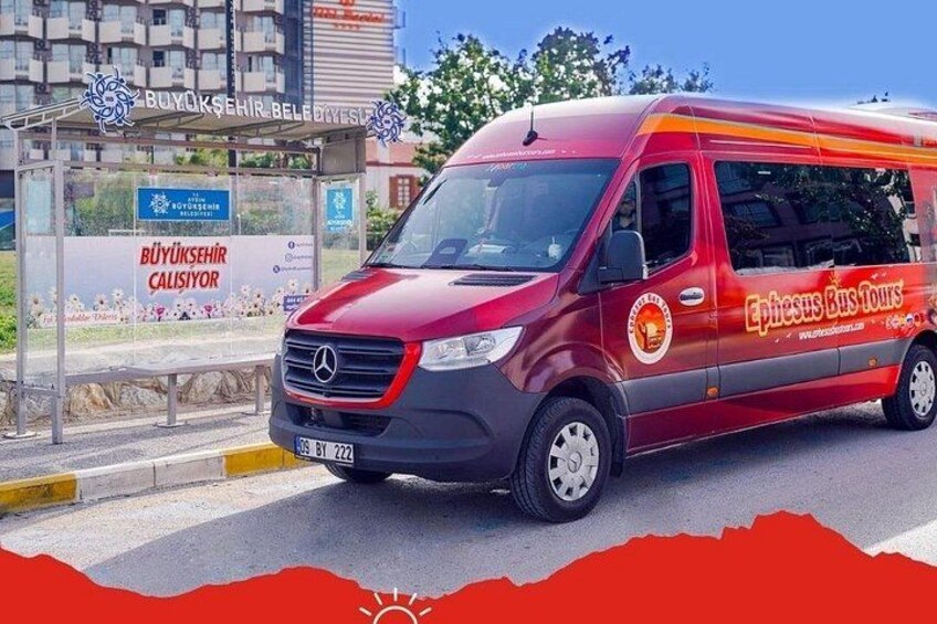 En röd buss med texten ”Ephesus Bus Tours” på sidan står parkerad på en gata. I bakgrunden syns en busshållplats och en byggnad. 1