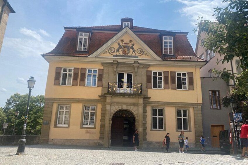 A historic building with a red-tiled roof, a central entrance, and a balcony with a coat of arms. 3
