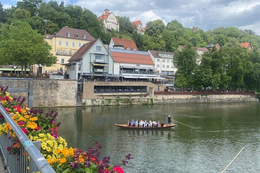 A river with a rowboat carrying people, a bridge with flowers, and buildings along the riverbank. 5