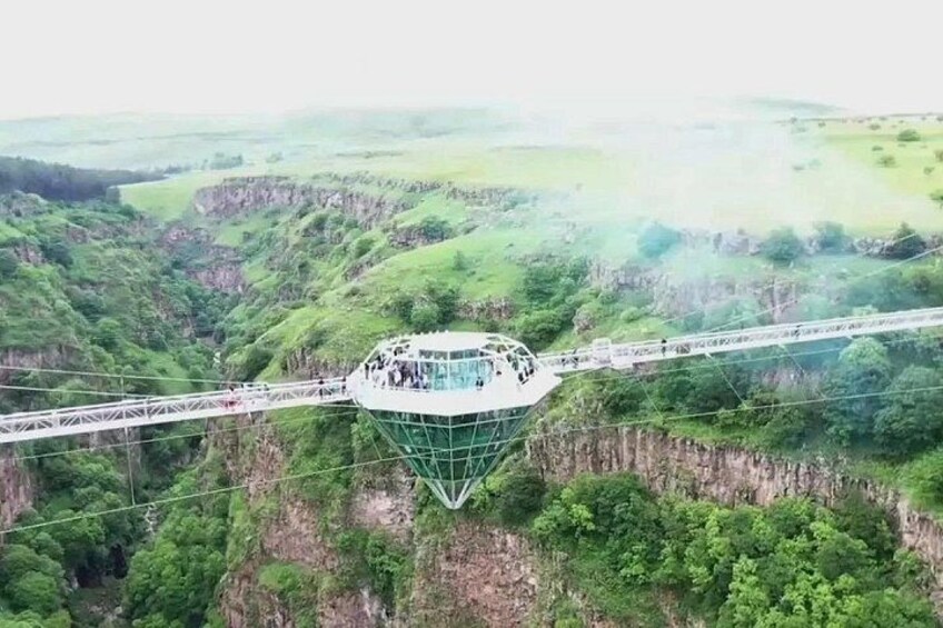 A large observation wheel with a glass floor, surrounded by lush green hills and a suspension bridge. 5