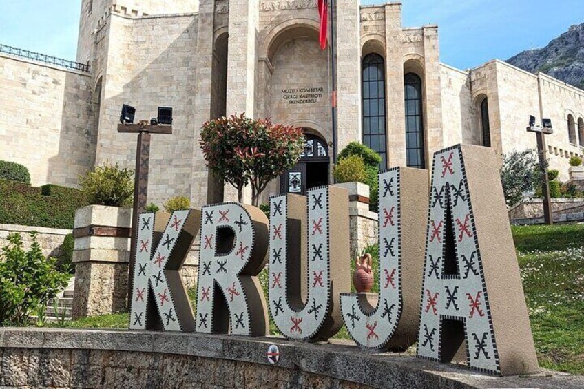 Grandi lettere con la scritta "KRUGA" a forma di croce, davanti a un edificio con finestre ad arco e una bandiera rossa. 1
