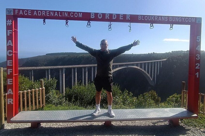 A person standing on a platform with arms outstretched, a red archway with text, and a bridge in the background. 1