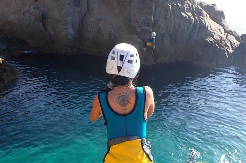 Una persona con camiseta azul y pantalones cortos amarillos, con casco blanco, de pie en agua azul cristalina y un acantilado rocoso al fondo. 2