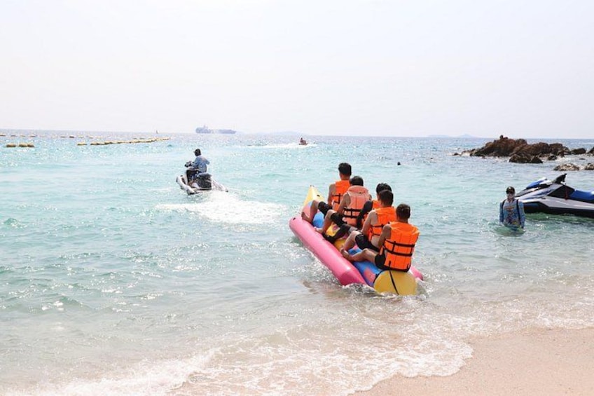 Un grupo de personas en un bote inflable rosa en aguas poco profundas, un jet ski al fondo y una costa rocosa. 5