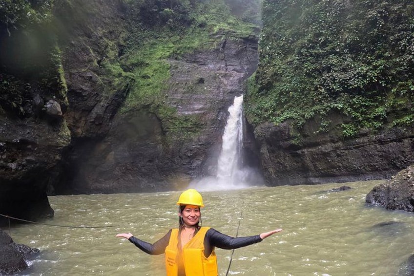 A person in a yellow life jacket and helmet standing in a river with a waterfall in the background. 2