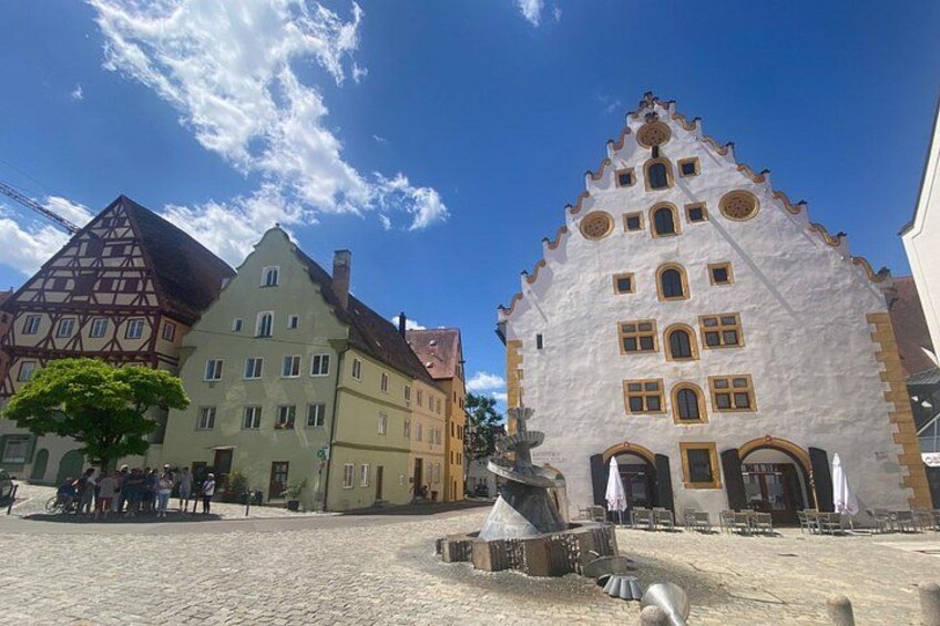 Una plaza adoquinada con edificios tradicionales europeos, una fuente y un cielo azul despejado. 5
