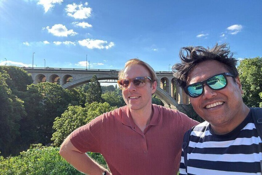 Two people standing in front of a bridge with greenery and a clear sky in the background. 3