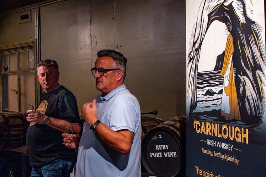 Two men standing in front of barrels, one holding a glass. A poster with a woman and the text 'CARNLOUGH Irish Whiskey' is visible. 4