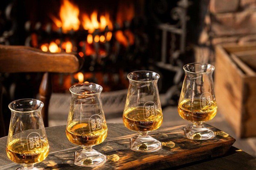 Four glasses of whiskey on a wooden tray with a fireplace in the background. 3