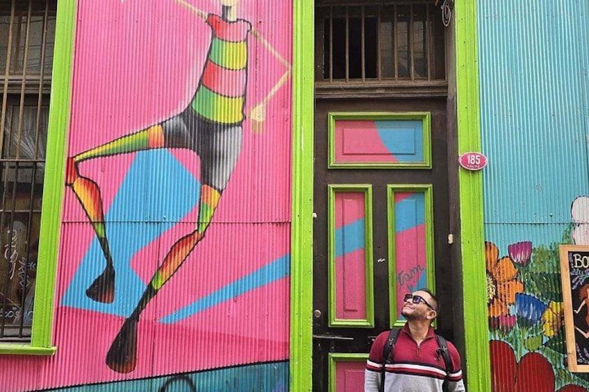 Un mural colorido en un edificio, una persona de pie frente a él, y una puerta con un cartel que indica el número 185. 2