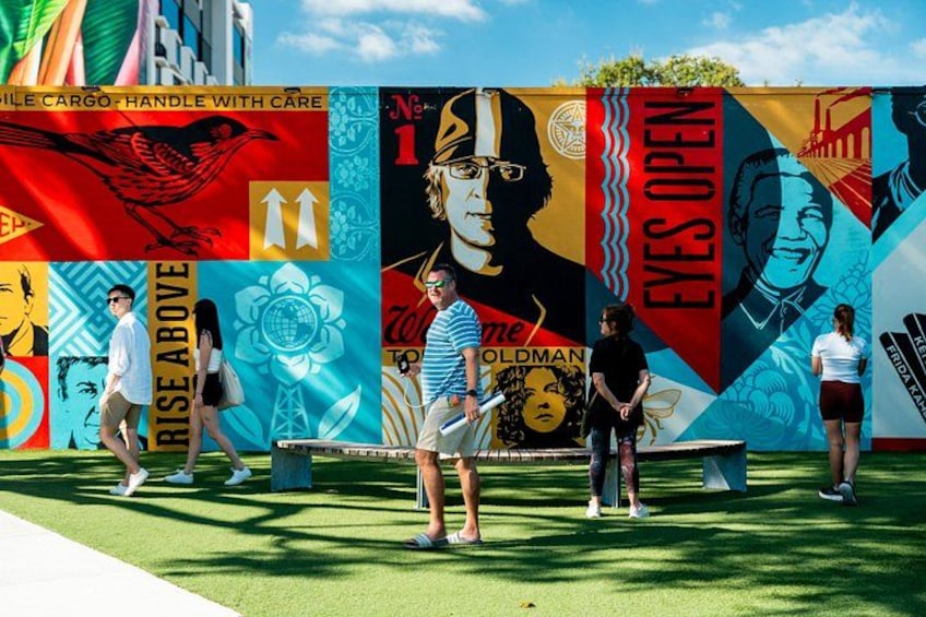 A mural with various figures and text, a green artificial grass area, and people walking. 1
