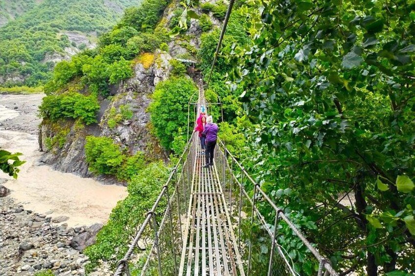 A person walking on a suspension bridge over a river, surrounded by greenery and rocky terrain. 3