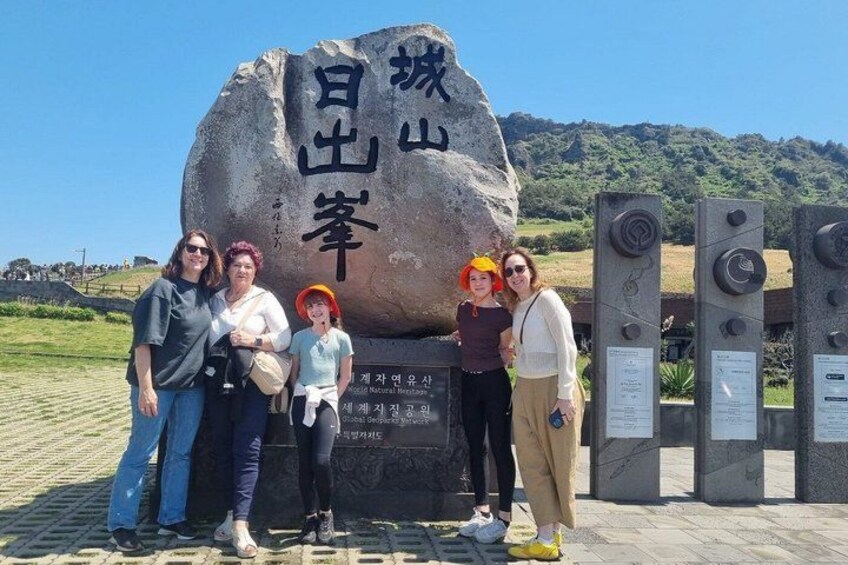 A large rock with Korean characters, a group of people posing in front, and informational plaques. 2
