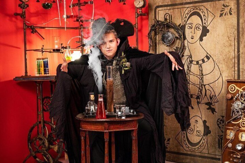 A person in a black outfit with a hat, sitting at a table with various scientific apparatus and a painting in the background. 2