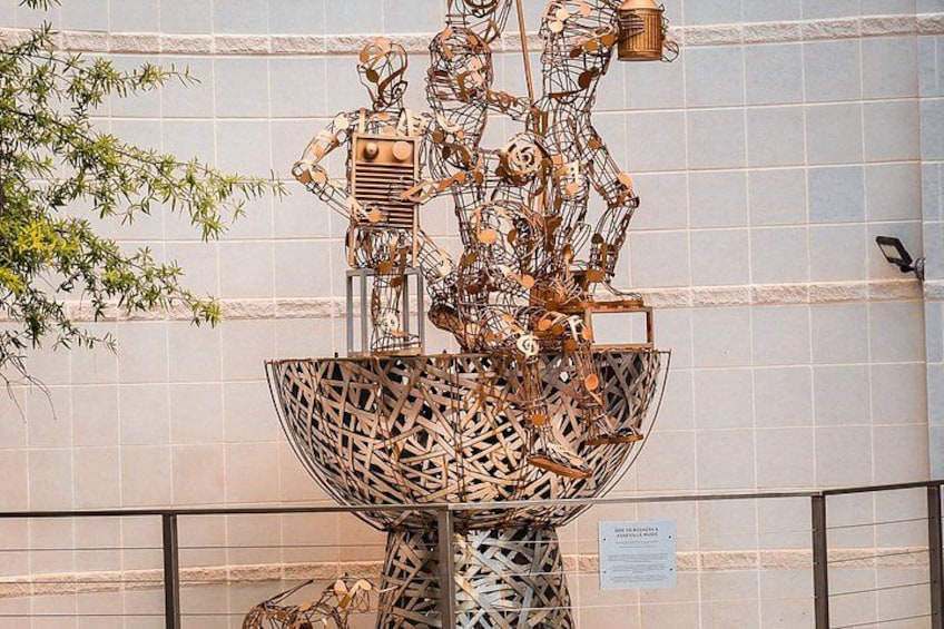 En stor metalskulptur med geometriske former og en kugleformet base, foran en flisebeklædt væg. 3
