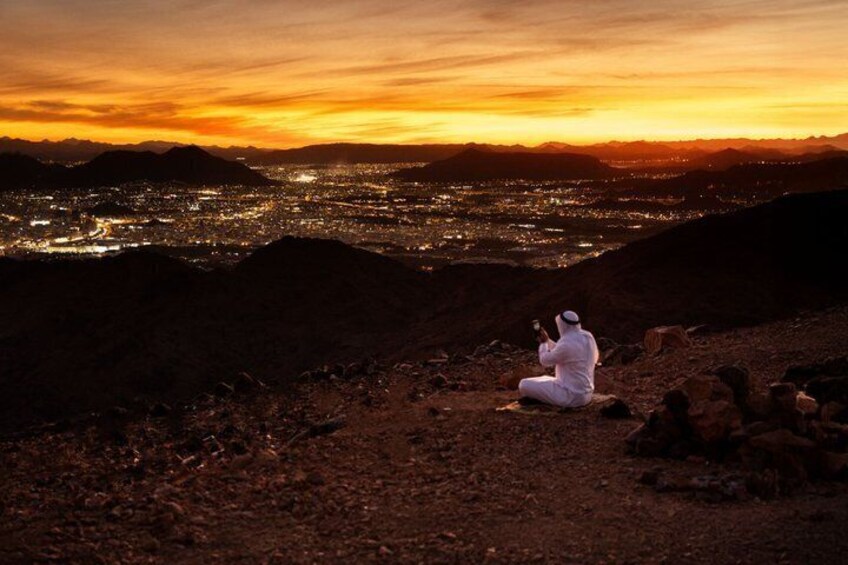 Eine Person in weißer Kleidung sitzt auf einem Felsen und blickt auf eine Stadtlandschaft bei Sonnenuntergang. 2